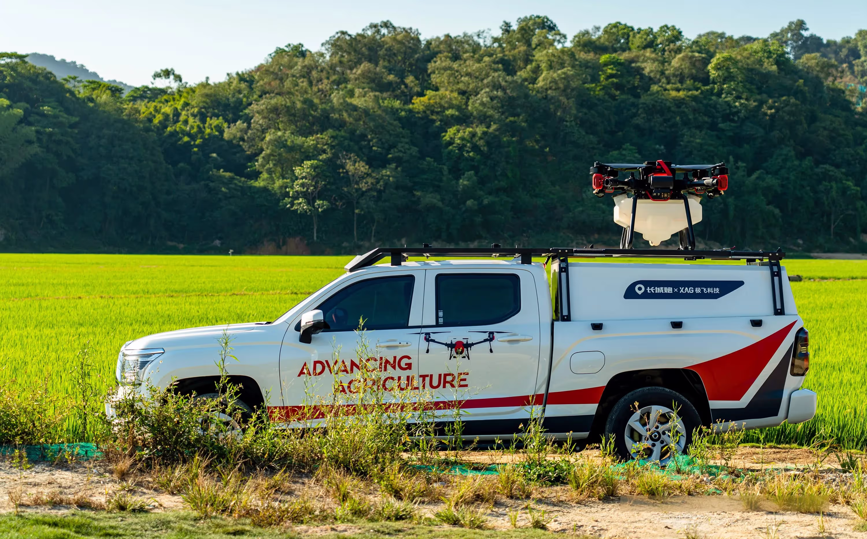 White pickup truck with 'Advancing Agriculture' text and drone mounted on its back in a green field.