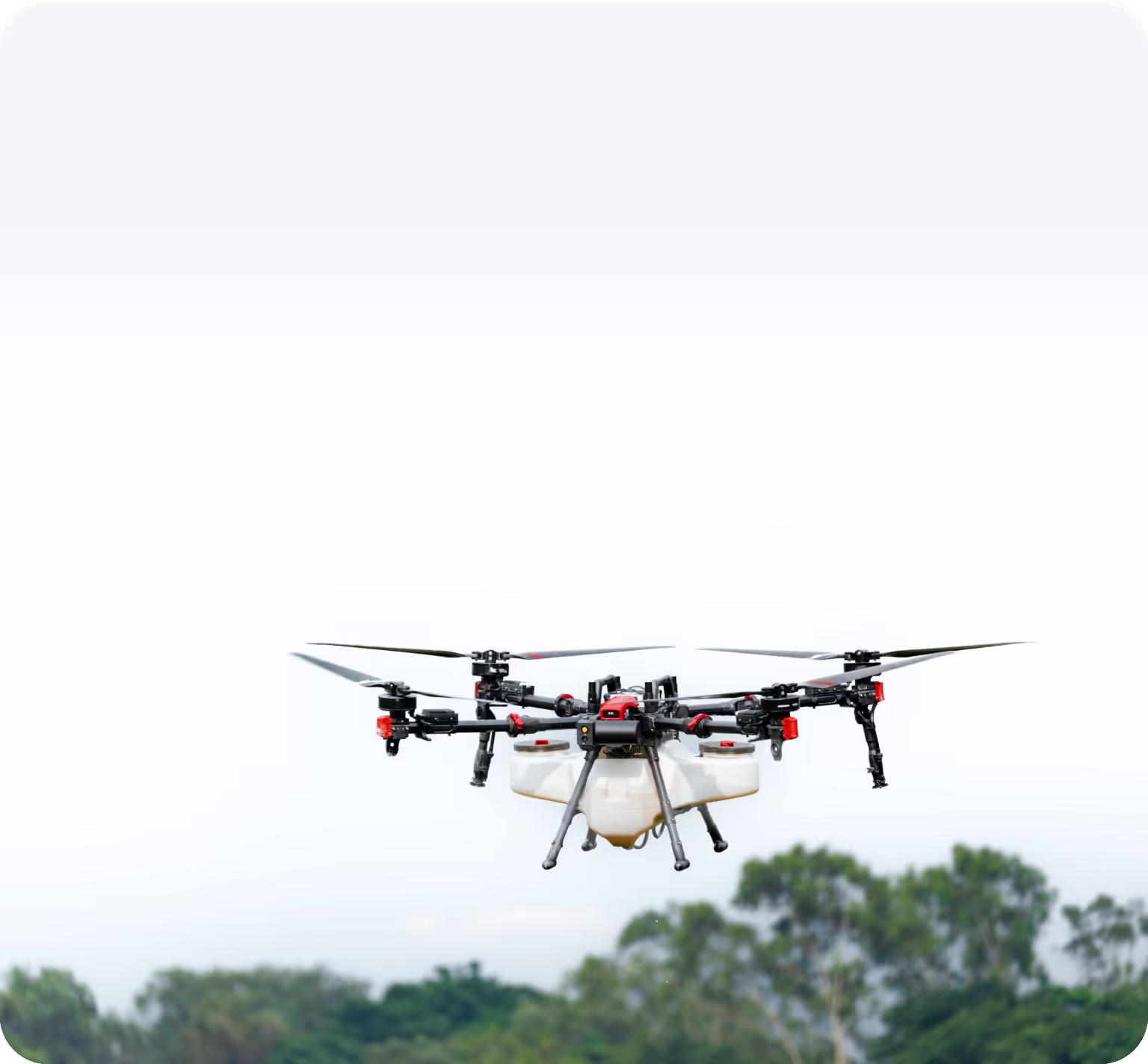 Agricultural drone flying outdoors above trees with a white payload tank and multiple rotors