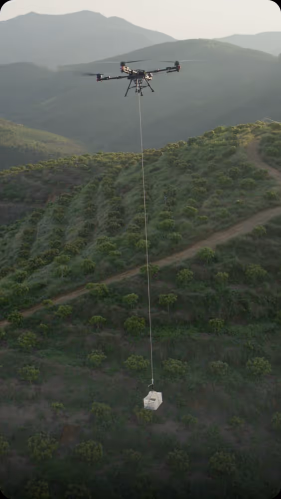 Drone flying over green hilly farmland carrying a white box suspended by a long cable.